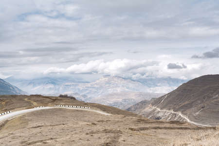 View of the alpine road against the background of the mountain peaks of a gloomy sky with clouds and dry grass of sand color. The concept of landscape, travel.の写真素材