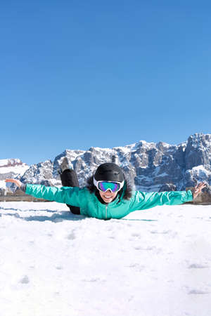 A teenage girl in a colorful ski suit, helmet and sunglasses lies in the snow and smiles happily against the backdrop of mountain peaks. Sports concept, people, travel.の写真素材