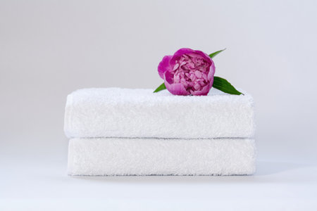 Two white neatly folded terry towels with a pink peony flower on a light backgroundの写真素材
