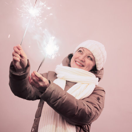 An adult woman in winter clothes holds sparkling sparklers in her hands against the background of a pink wall.の写真素材