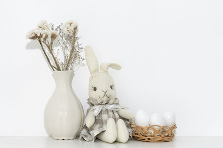 Easter composition with a soft rabbit igushka, white eggs in a basket and a vase of flowers on a shelf in the roomの写真素材
