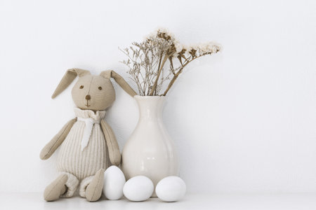 Easter composition with a soft rabbit igushka, white eggs in a basket and a vase of flowers on a shelf in the roomの写真素材