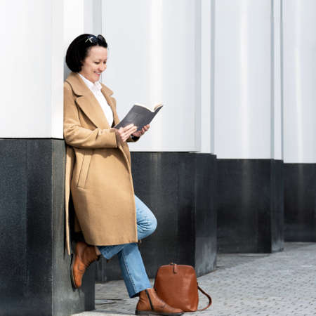 An adult woman fashionably dressed stands on the street near a building reading a book.の写真素材