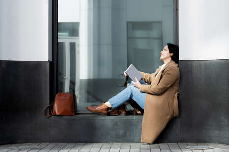 An adult woman fashionably dressed sits on the street on the windowsill of a building reading a book.の写真素材