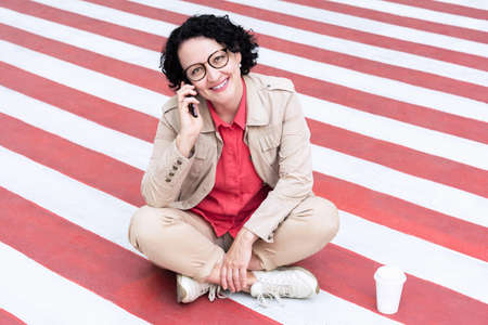 An adult woman with glasses with black hair sits on the sidewalk talking on the phone with a smile on her face..の写真素材