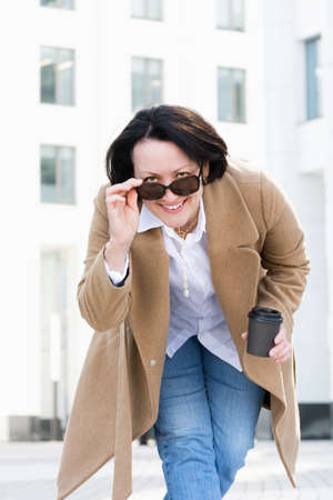 woman stands on the street with a paper cup of coffee looks out from under sunglasses and smiles.の写真素材