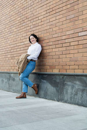 A white-skinned woman in fashionable clothes stands against the backdrop of a red brick building.の写真素材