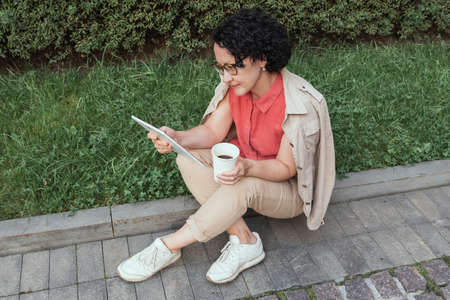 An adult woman sits on the lawn, drinks coffee and looks into a tablet.の写真素材