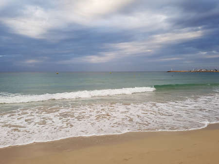 View of the sea coast with a sandy beach, the ocean with waves and a lighthouse in the distance against the backdrop of a gloomy sky..の写真素材