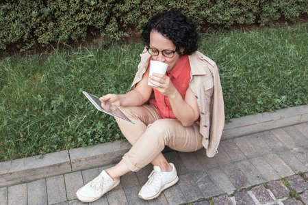 An adult woman sits on the lawn, drinks coffee and looks into a tabletの写真素材
