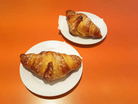 Two freshly baked croissants lie on white saucers on an orange tableの写真素材