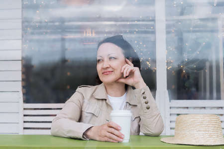 Happy mature woman with a glass of coffee sits at a table on the summer veranda of a cafe and smiles.の写真素材