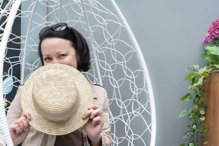 Smiling woman sits in an armchair on the summer terrace of a cafe and covers her face with a straw hat.の写真素材