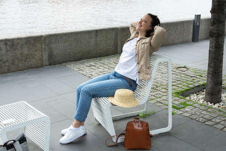 Happy young woman is resting on a bench on the embankment of the river in summer and smiling.の写真素材