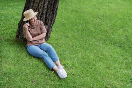 A woman in summer clothes and a straw hat sits near a tree on the lawn and smiles.の写真素材