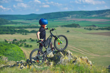 Little child stand next to his mountain bike on mountains edge and looks at the beautiful sceneryの写真素材