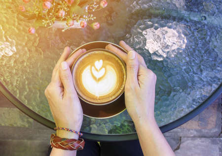 Hands with bracelets on wrist  holding cup of hot coffee latte cappuccino on round glass top table, filtered color effect.の写真素材