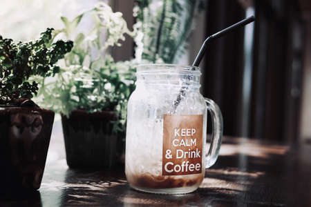 Relaxing atmosphere in a coffee shop, glass of iced cold coffee with trendy quote "Keep calm & drink coffee" and blurred small potted green plants in background. Summer refreshing drinks.の写真素材
