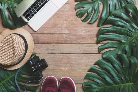 Flat lay creative frame of tropical nature leaves Monstera on rustic wood grunge background with laptop computer, retro camera, hat and pair of sneaker shoes, digital nomad and life balance concepts.の写真素材