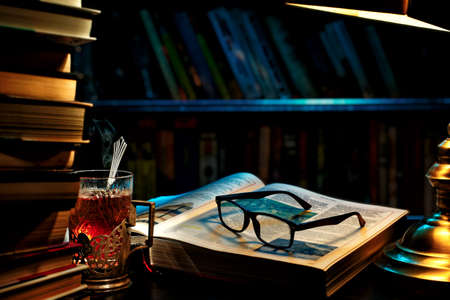 library. A book unfolded under a desk lamp, with simple black glasses on it. A glass of hot tea. Background of the book on the shelves.の写真素材