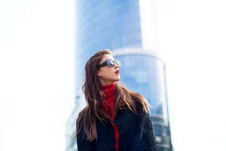 Fashion portrait of young elegant confident woman outdoor. black jacket, red lips and sunglasses. She looking at the sky On the background of the big city.の写真素材