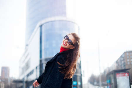 Cool girl with long hairstyle and red lips having fun in the city. She wears sunglasses and smiling to camera with snow-white smile. On the background of the big city.の写真素材