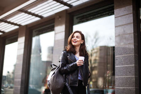 The concept of street fashion. portrait of Young girl dressed in a fashionable outfit. Posing against the window of the boutique girl smiling and drinking coffeeの写真素材