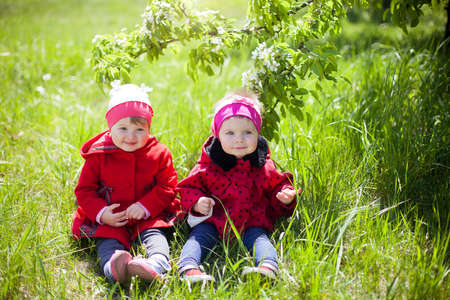 Two little bridesmaid sitting on the grass and smiling .Portrait of two little girls in red coats in the Park on beautiful sunny dayの写真素材