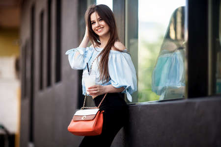 portrait of Young girl dressed in a fashionable outfit. Posing against the window of the boutique girl smiling and drinking coffeeの写真素材