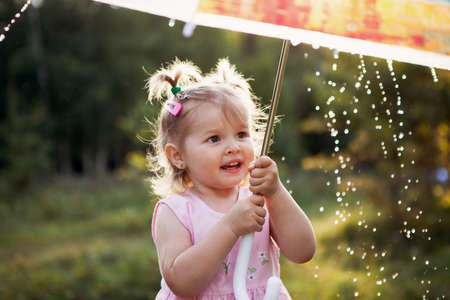 small cheerful girl holds a transparent umbrella and looks at the raindrops. The weather. Cute baby in the rainの写真素材