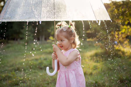 toddler girl standing on the nature outdoors holding umbrella on a rainy summer day.girl is holding a large umbrella. The drops are flowing down the umbrella.の写真素材