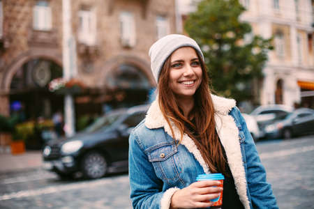 Portrait pretty girl with long hair in denim coat walking on street with coffee. She looks excited...