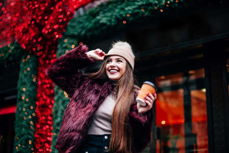 The concept of street fashion. portrait of Young girl dressed in a fashionable outfit. Posing against the window of the boutique girl smiling and drinking coffee .の写真素材