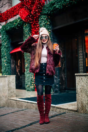 Portrait pretty girl with long hair in Burgundy fur coat of artificial fur knitted hat and bright burgundy lips walking on street with coffee to go. She looks excited to camera City lifestyle .の写真素材