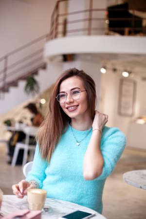 young stylish beautiful woman sitting in city cafe in street, spring fashion trend style, drinking latte, long hair, reading diary, smiling, happyの写真素材