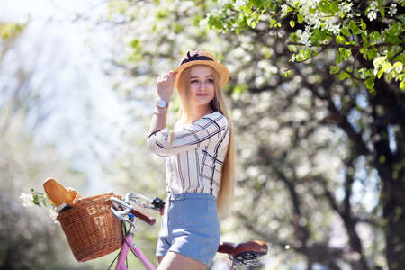 beautiful blonde girl holds a bicycle with a basket of flowers, and smiles a sweet smile. the girl...