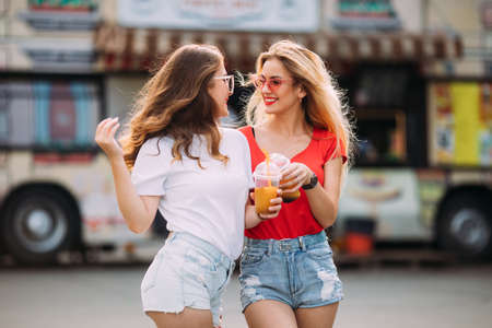 two young stylish girls talk and laugh. hold cocktails in hands. Holiday in the park going crazy together, wearing mirrored sunglasses, amazing view on the city, bright colors evening sunlight.の写真素材