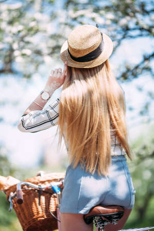 Close-up portrait of cheerful woman wearing straw hat sitting on pink retro bicycle, on blur background. Photo of fashionable girl with beautiful long hair back to camera.の写真素材