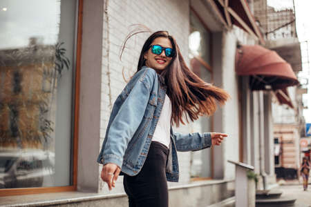 Young beautiful model girl in sunglasses smiles and looks back in the city. Dynamically young girl walks down the street. Hair fluttering in the windの写真素材