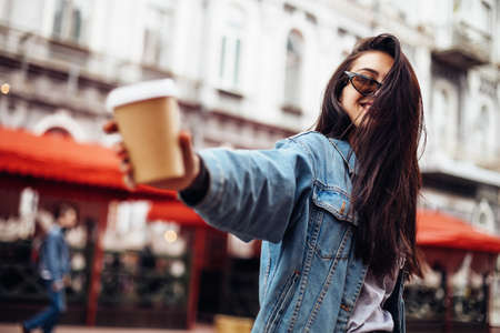 Stylish happy young woman wearing woman in a denim jacket. She holds coffee to go. portrait of smiling girl in sunglasses and bag. Emotional womanの写真素材