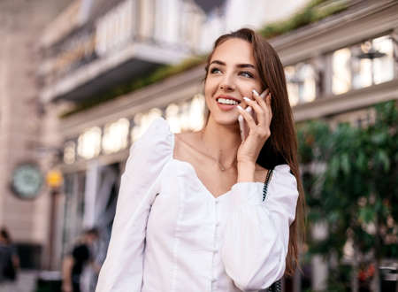 Portrait attractive young girl dressed in white shirt talking on phone in city street. stylish girl...