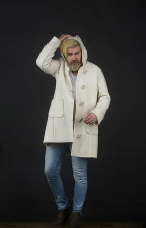 Elegant bearded man in coat puts on a hood, standing on a dark background.