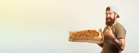 Happy hungry bearded man eating pizza and showing thumbs up gesture isolated on white background.の写真素材