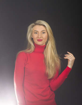 Optimistic girl touches her blond hair. Lady in red blouse on dark background.