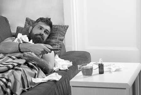 Bearded ill man lying in bed at home. Sick man lying on sofa with TV remote under a blanket at home...