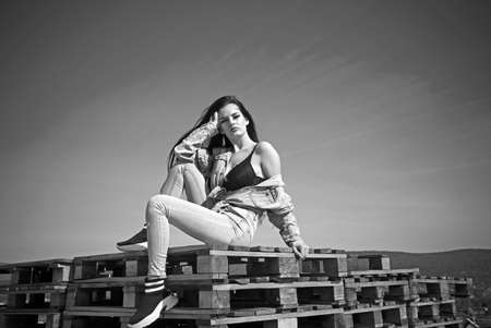 Beautiful sexy girl with long legs in jeans short overalls sits on pallets setting her long silky...