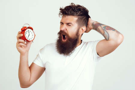 Handsome man yawning holds vintage alarm clock over isolated background. Hipster with tattoo gets up...
