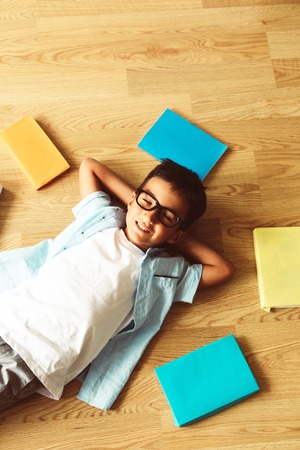 Smart preschool boy in blue shirt with eye glasses smiling, sitting on the floor with colorful books and pens, looking to the camera, home schooling and back to school, wunderkind conceptの写真素材