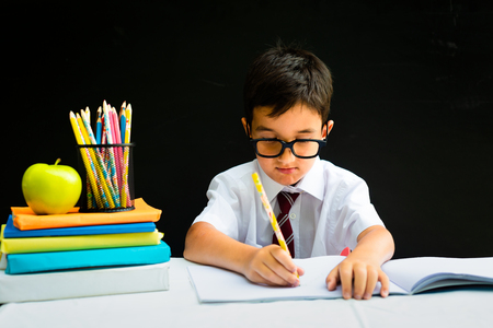 Cute smart handsome school boy in white shirt with eye glasses, writing at the desk in classroom, happy first grader pupil, smiling, knowledge study black board in the background, back to school conceptの写真素材