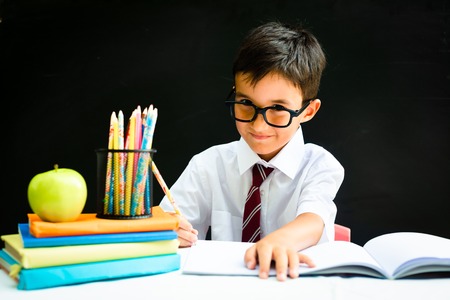 Cute smart handsome school boy in white shirt with eye glasses, writing at the desk in classroom, happy first grader pupil, smiling, knowledge study black board in the background, back to school conceptの写真素材